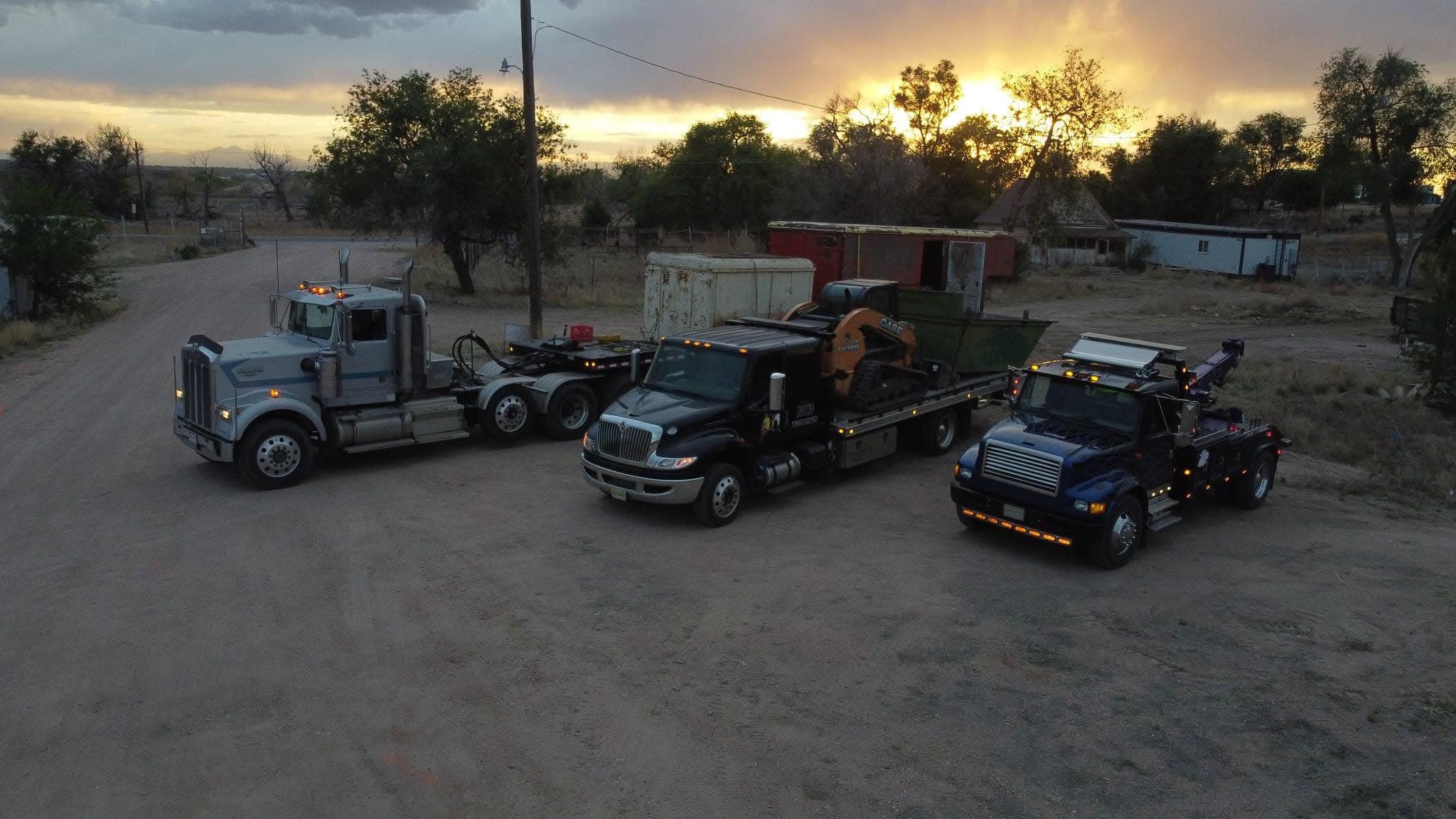 Chuck's Towing fleet lined up at sunset — semi tractor, flatbed with skid steer, and heavy wrecker